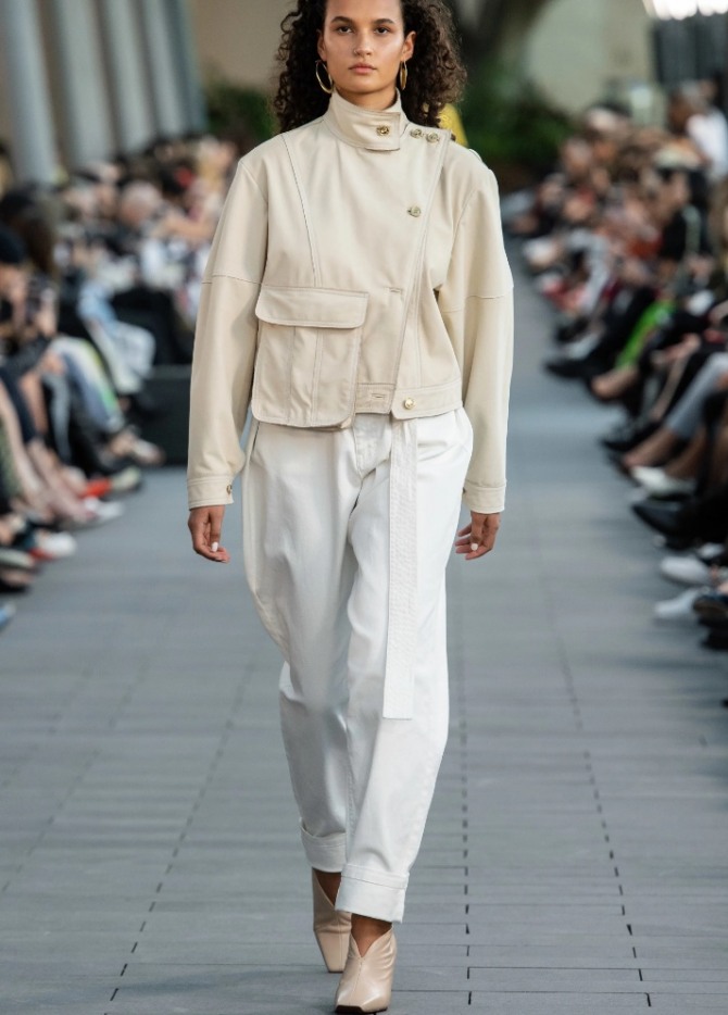красивый и стильный уличный образ в светлых тонах на весну-лето 2020 года - белые брюки с курткой, имеющей воротник-стойку и один большой накладной карман на груди, с асимметричной косой застежкой на молнию - фото с модных показов Australia Resort 2020